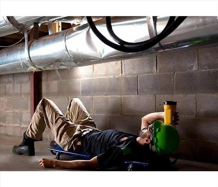 SERVPRO technician in a basement laying under a ventilation pipe examining it with a flashlight
